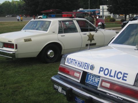 1991 Ford LTD Crown Victoria - cream sheriff sedan with a gold star decal sits behind the rear of a black-and-white North Haven Police Plymouth.