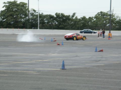 2001 Mercury Grand Marquis - A sports car performs a donut maneuver on a paved lot, kicking up smoke, with other vehicles and spectators in the background.