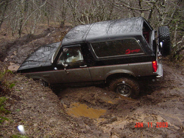 Two-tone Ford Bronco tilted in a deep mud rut on a forest trail, articulating front wheel