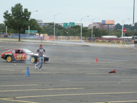2001 Mercury Grand Marquis - A sports car with racing decals drifts in a parking lot, with a person walking nearby and cones marking a course.