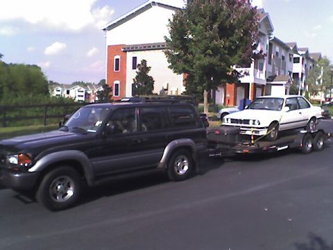 1997 Toyota Land Cruiser - 1997 Toyota Land Cruiser towing a white BMW E30 on a trailer down a street.