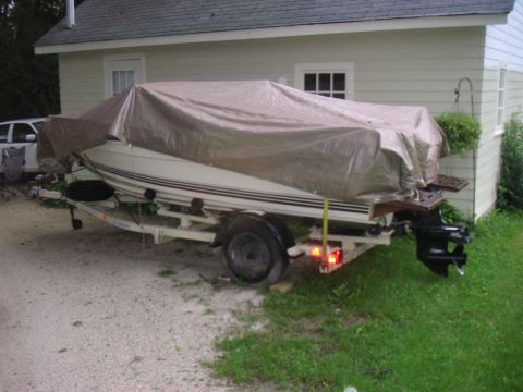 1979 Four Winns 170 Horizon - A boat on a trailer is covered by a tarp, parked on a gravel driveway next to a house.