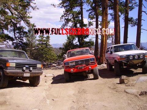 1990 Ford Bronco - Three modified SUVs, including a 1990 Ford Bronco, are parked on a dirt road in a wooded, mountainous area.