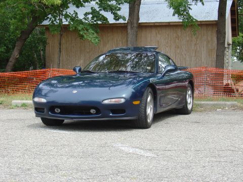 1986 Mazda RX-7 - A dark blue Mazda RX-7 is parked in front of a wooden building, with orange safety netting in the background.