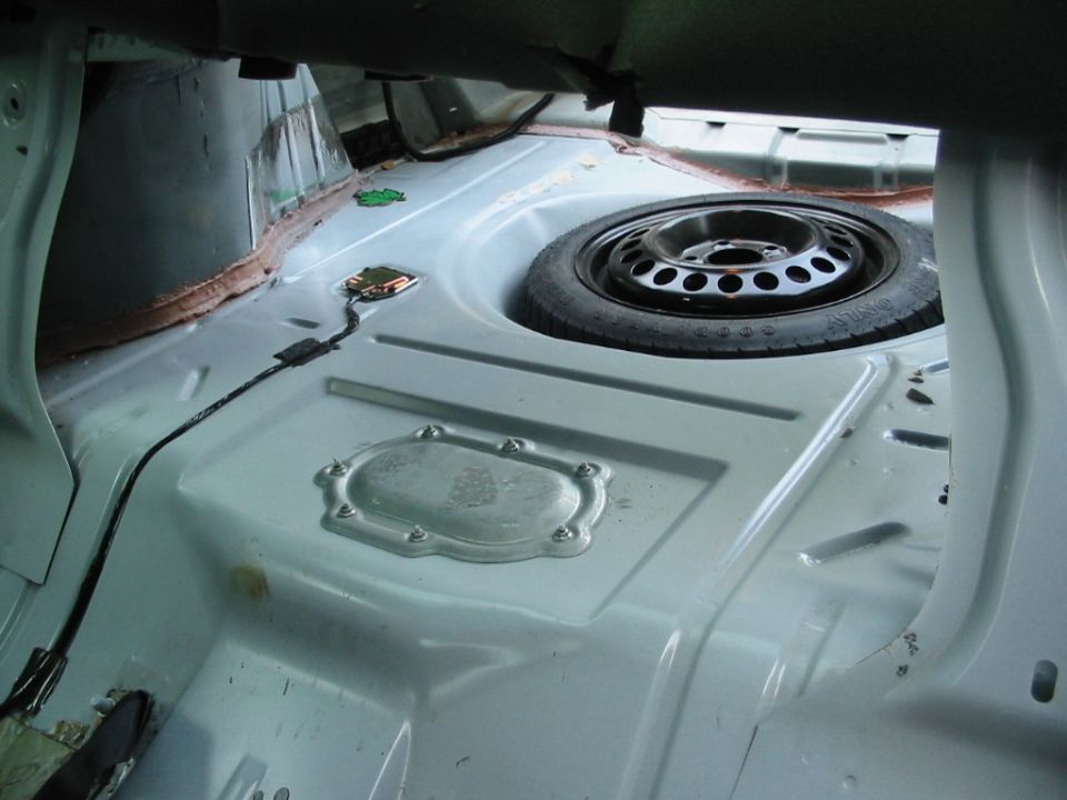 Stripped car trunk interior showing bare painted metal floor, compact spare tire, access cover, and wiring.