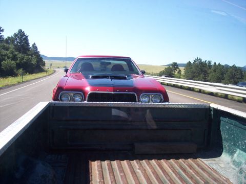 1972 Ford Ranchero - A red 1972 Ford Ranchero with a black hood scoop drives down a highway, seen from the bed of a pickup truck.