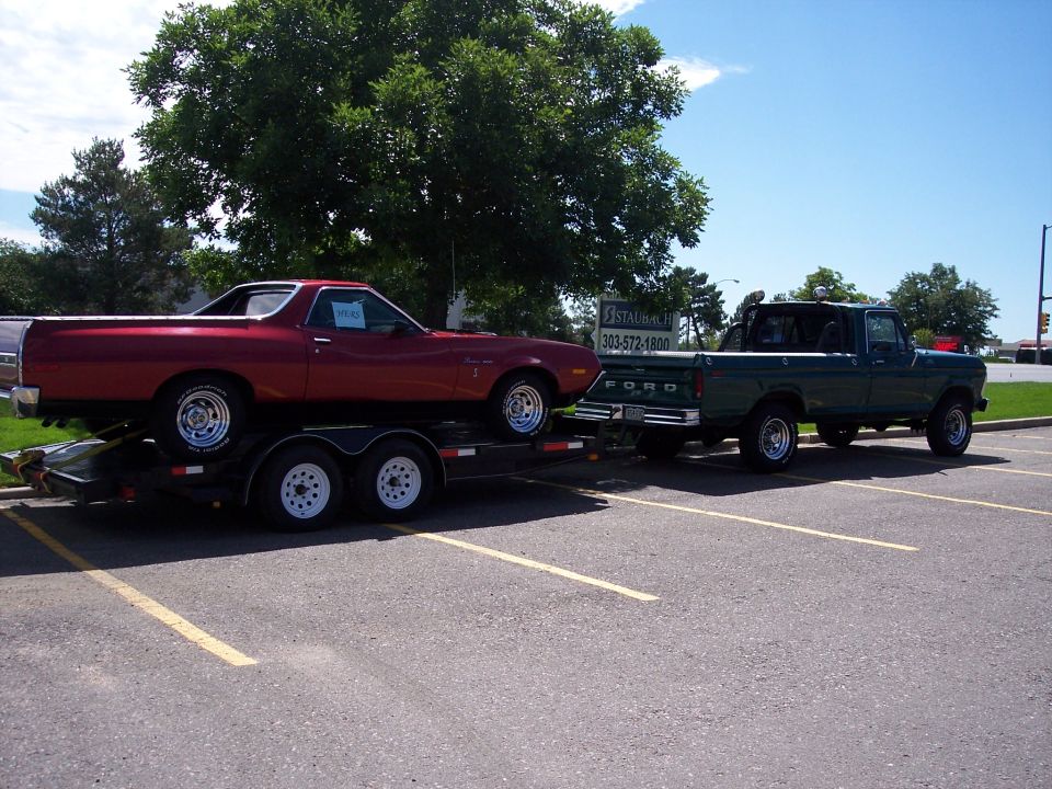 Maroon Ford Ranchero on a trailer towed by a green Ford pickup in a parking lot