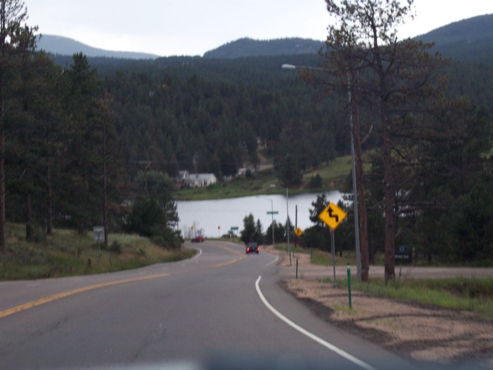 Curving mountain road heading down toward a lake with forested hills and a yellow curve warning sign