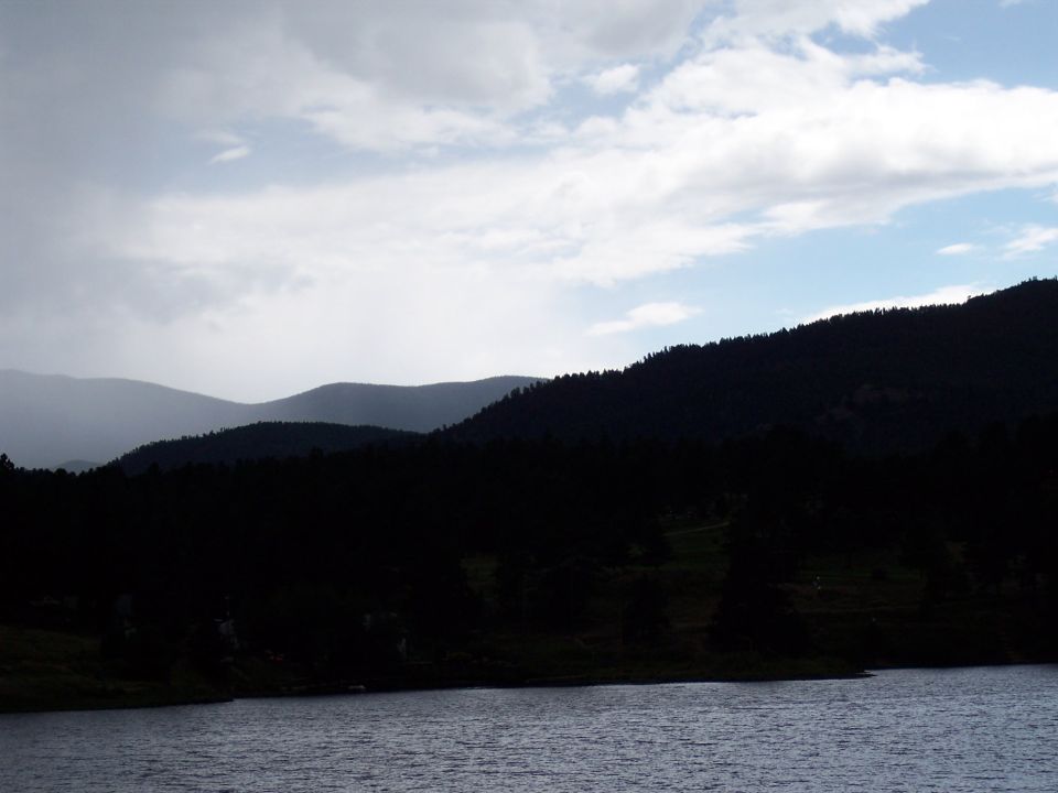 Forested mountains under cloudy sky above a dark lake