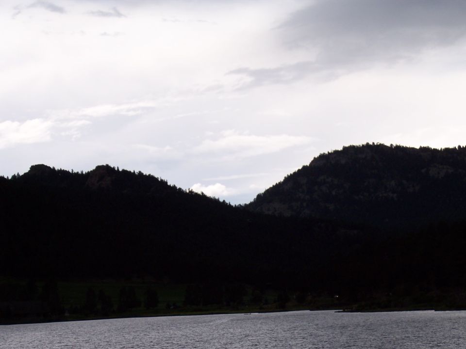Silhouetted forested mountains under a cloudy sky above a lake.