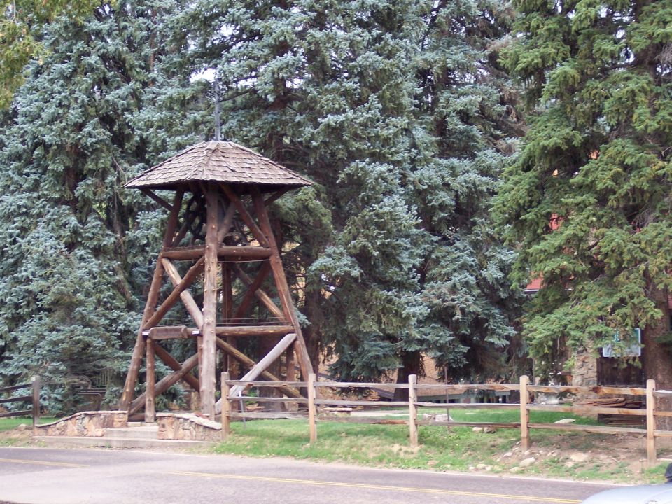 Wooden bell tower with shingle roof behind a split-rail fence among pine trees