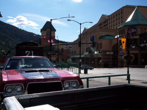 1972 Ford Ranchero - A red coupe with a scoop on its hood is parked on a street in a town with mountains and large buildings.