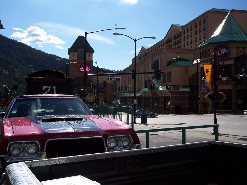 Maroon Ford with dual headlights and hood scoop parked on a town street with casino buildings behind.