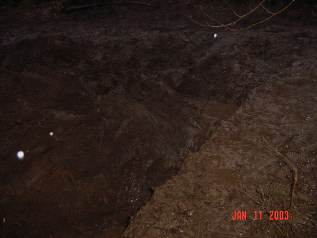 Black-and-tan Ford Bronco stuck in a deep mud hole with three onlookers standing nearby