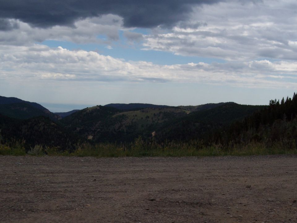 Forested mountain ridges under cloudy sky, gravel foreground