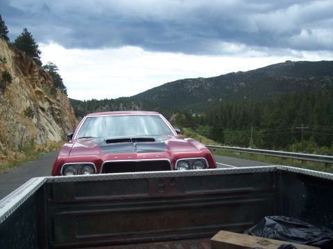 1972 Ford Ranchero - A red coupe with a black hood scoop is seen from the bed of a pickup truck on a scenic mountain road.