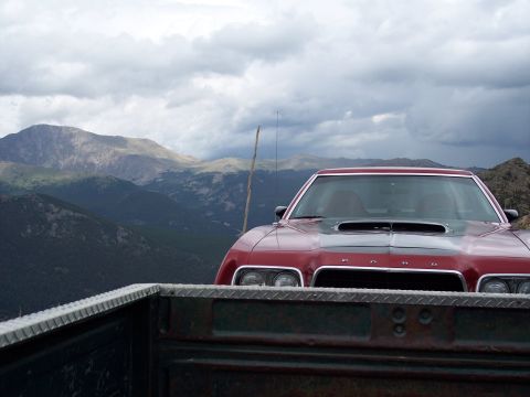 1972 Ford Ranchero - A red car with a prominent hood scoop is seen from the perspective of a pickup truck bed, with mountains under a cloudy sky.