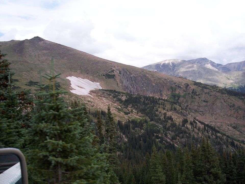 Mountain view with pines and snow patch, seen past a vehicle's side mirror