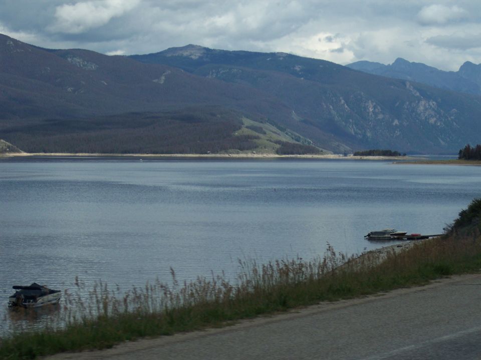 Overcast mountain lake with distant boats and a roadside shore