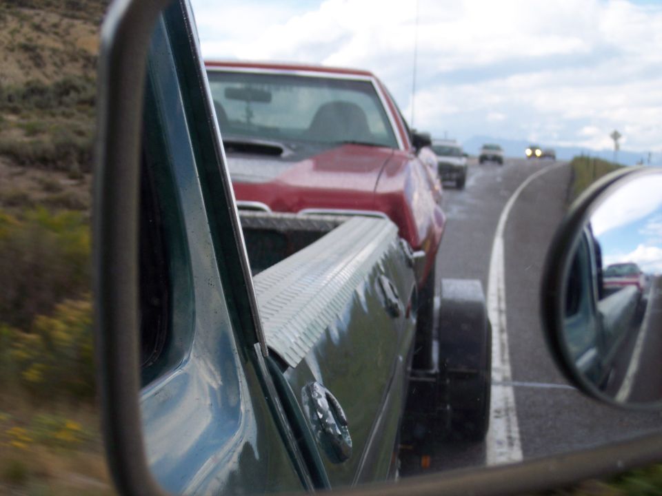 View past a green car's hood and side mirror toward a red car on a rural highway