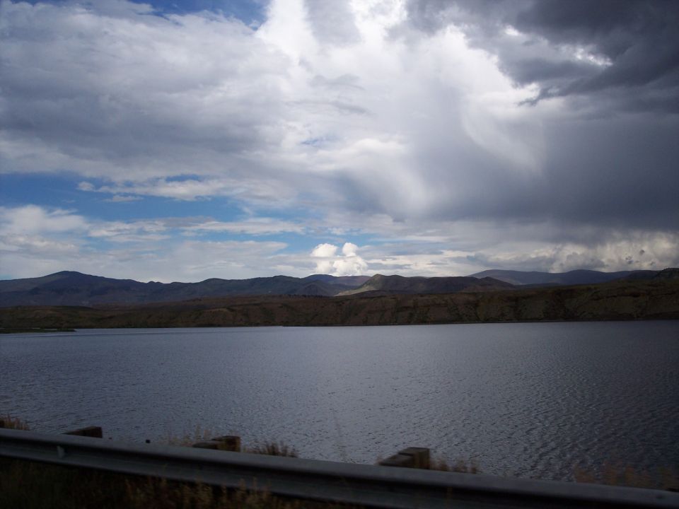 Lake with brown hills under cloudy sky, seen past a metal guardrail