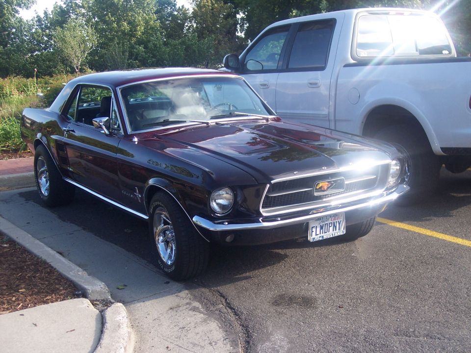 Dark burgundy vintage Ford Mustang coupe parked, 3/4 front view, white pickup beside it.