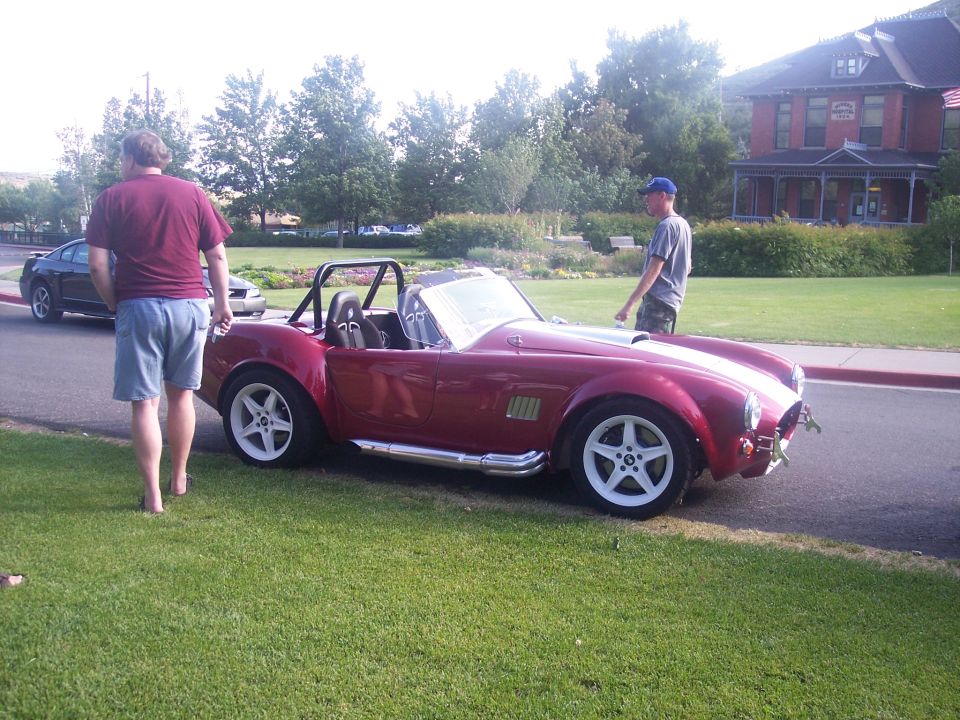 Red Cobra-style roadster with white stripe parked by a lawn with two men nearby