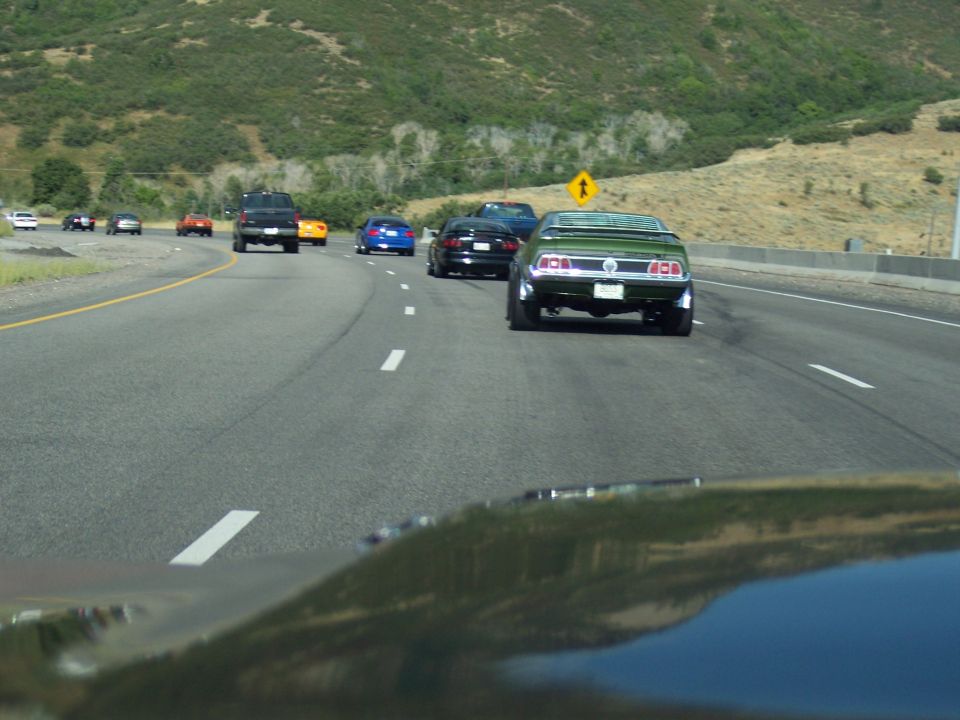 Dark green classic Mustang fastback seen from behind on a highway with other traffic and hills.