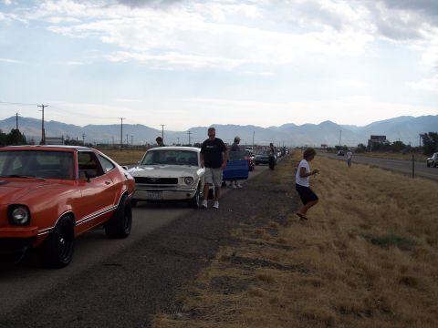 1972 Ford Ranchero - A line of cars, including an orange vehicle with black wheels and a white car, are parked on the side of a road with mountains in the backg…