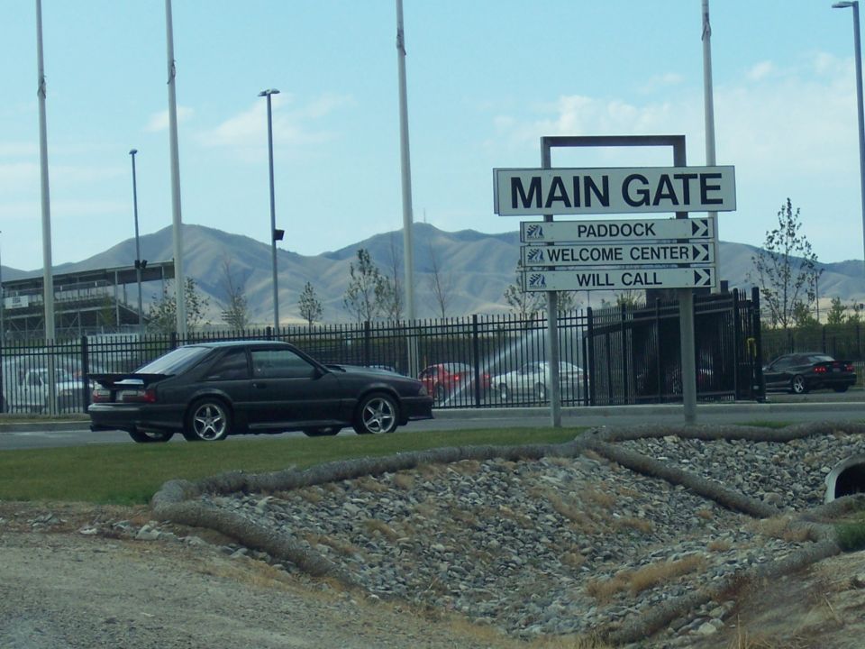 Dark Fox-body Mustang hatchback driving past a fence and 'MAIN GATE' sign