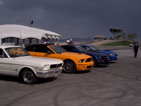 1972 Ford Ranchero - A lineup of Ford Mustangs, including a classic 1965 model and newer generations, are parked on asphalt under a cloudy sky.