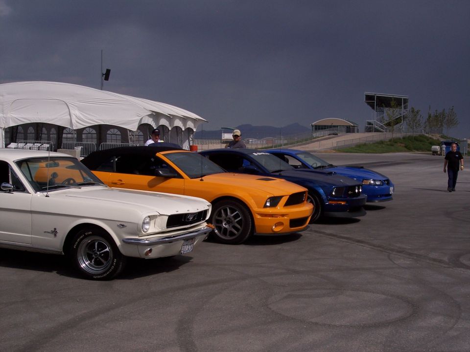 Row of Mustangs (white classic, orange, blue) parked on pavement under stormy sky.