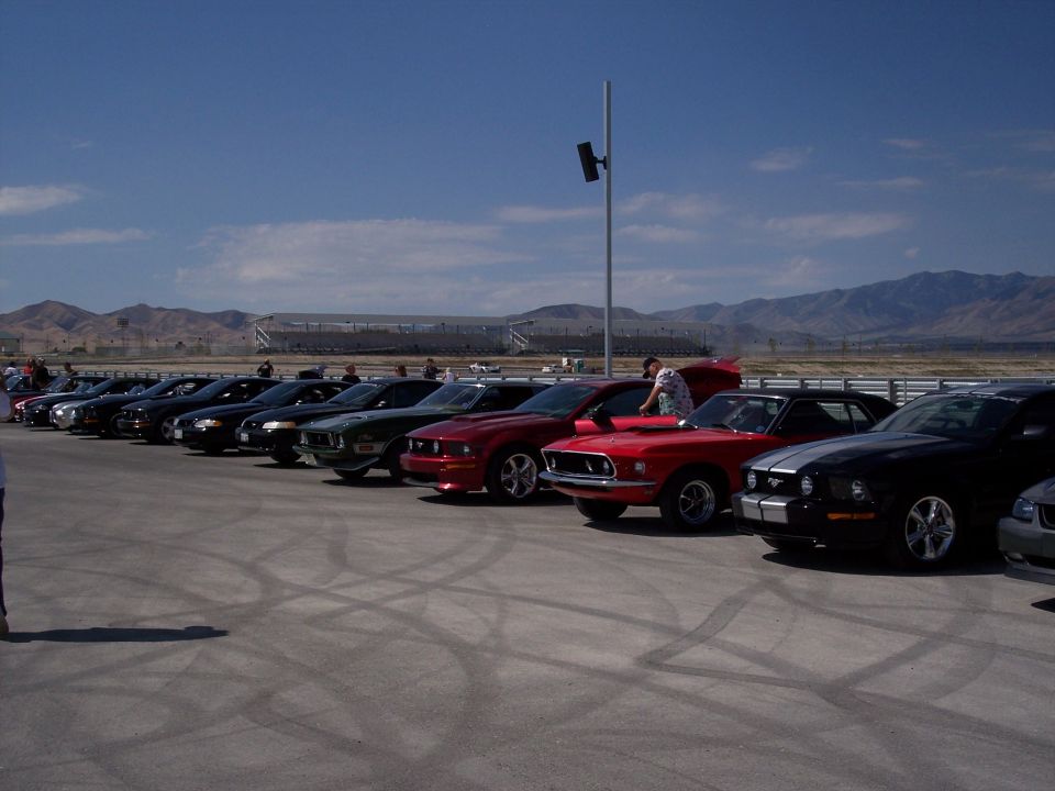 Row of parked Mustangs including red and black cars at a racetrack, mountains behind.