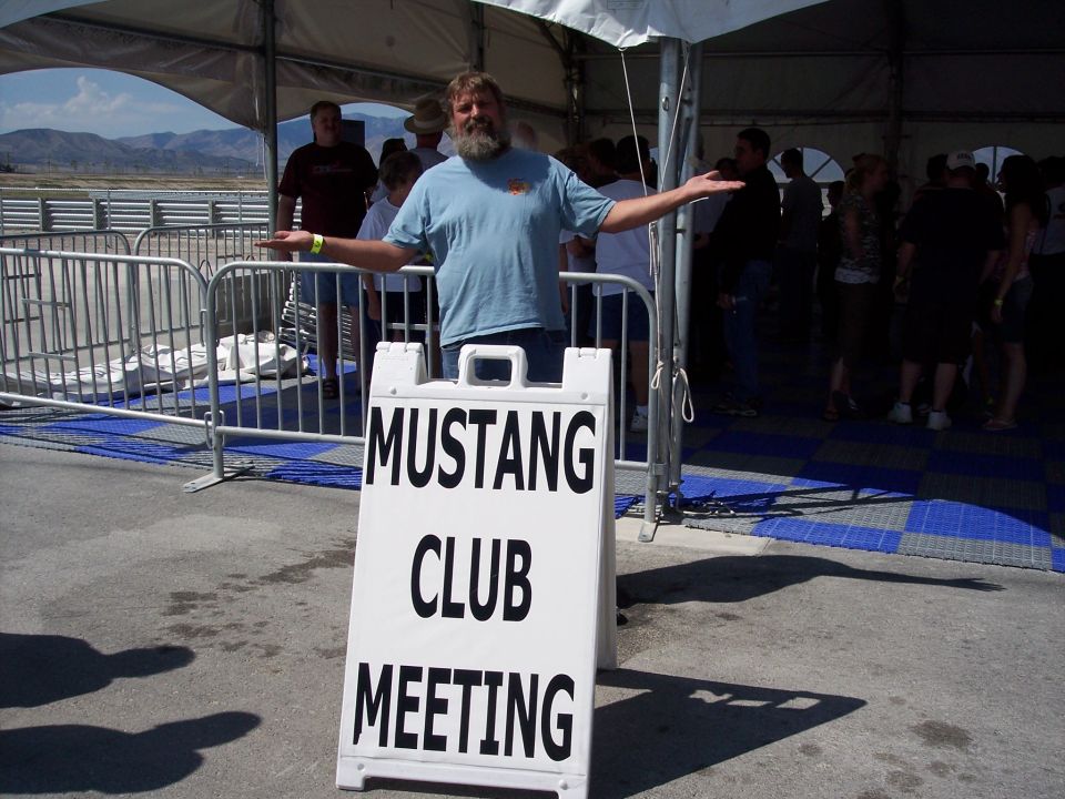 Man gesturing behind a 'Mustang Club Meeting' sign under an event tent