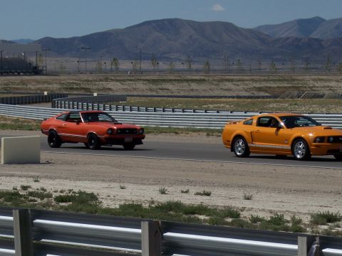 1972 Ford Ranchero - Two Ford Mustangs, one older model and one newer, race on a track with mountains in the background.