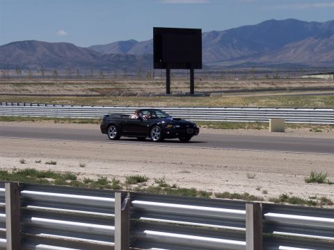 1972 Ford Ranchero - A black convertible sports car cruises down a racetrack under a clear sky, with a large digital billboard visible in the distance.