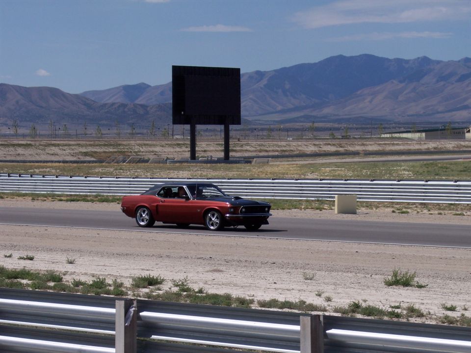 Red coupe with black hood and chrome wheels driving on a racetrack with mountains behind.