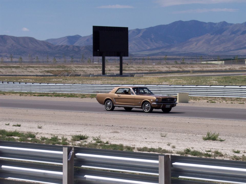 Tan vintage Ford Mustang coupe on a racetrack with mountains behind