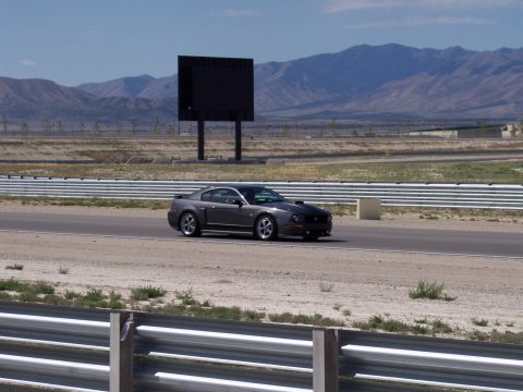 1972 Ford Ranchero - A dark gray coupe, likely a 2005 Ford Mustang, drives on a racetrack with mountains in the background.