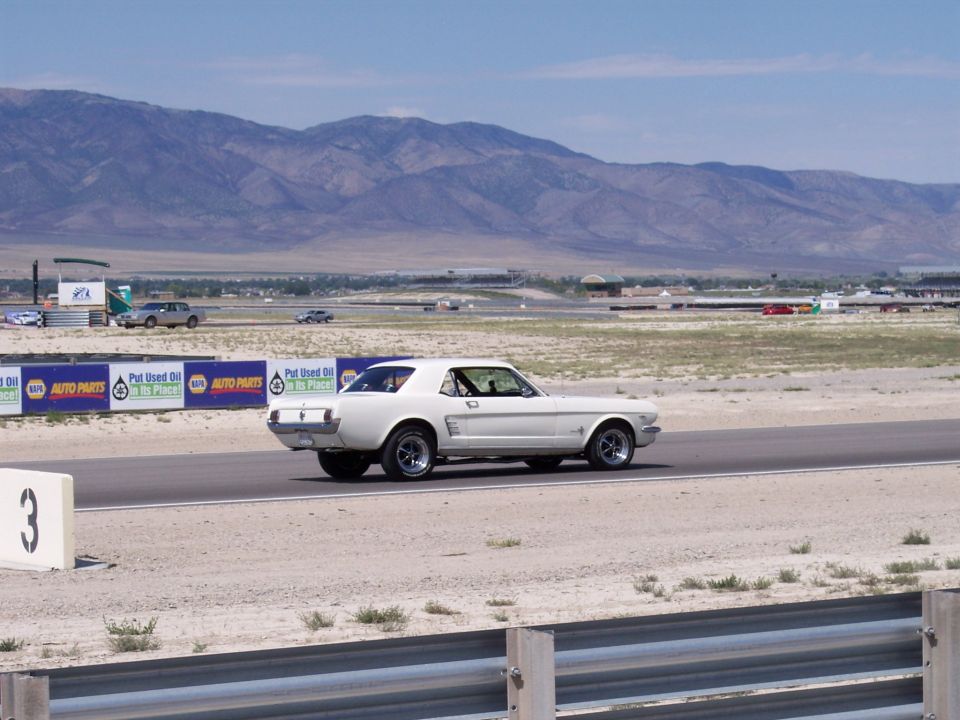 White vintage Ford Mustang coupe on a racetrack, side view, with sponsor banners and mountains behind.