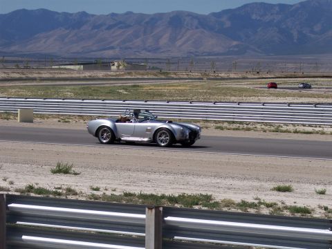 1972 Ford Ranchero - A silver Shelby Cobra speeds around a race track under a clear sky with mountains in the distance.