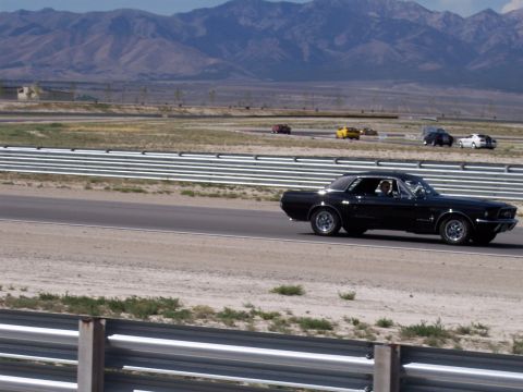 1972 Ford Ranchero - A black coupe drives on a race track with other cars in the distance and mountains behind.