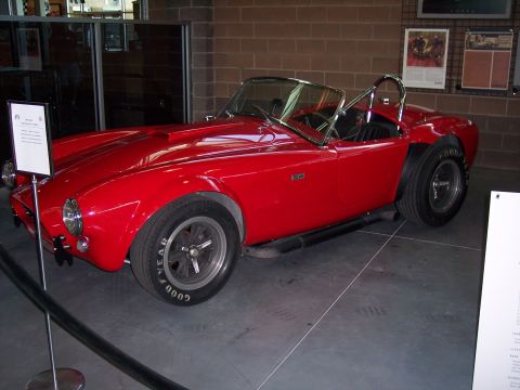 1972 Ford Ranchero - A red convertible sports car with a distinctive hood scoop and side pipes is displayed indoors.