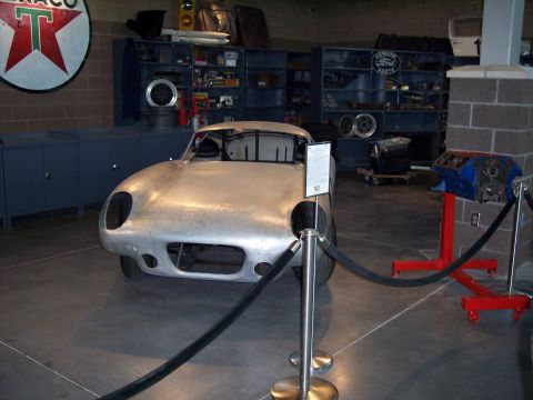 1972 Ford Ranchero - A bare aluminum sports car sits on display in a garage-like setting with shelves of parts in the background.