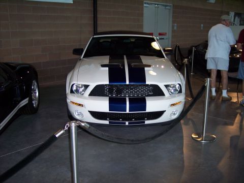 1972 Ford Ranchero - A white 2005 Ford Mustang GT500 with blue racing stripes is displayed indoors, with another car visible to the left.