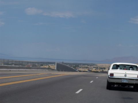 1972 Ford Ranchero - A white 1965 Ford Mustang coupe drives on a highway with a distant city and mountains in the background.