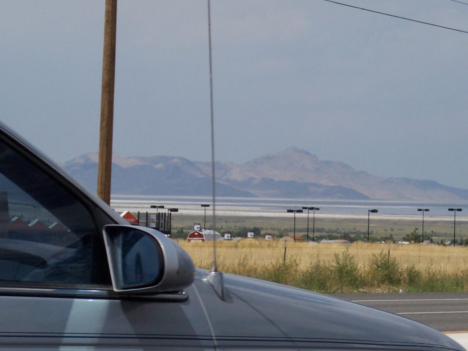 Silver vehicle's front corner with side mirror and antenna, mountains and field in background