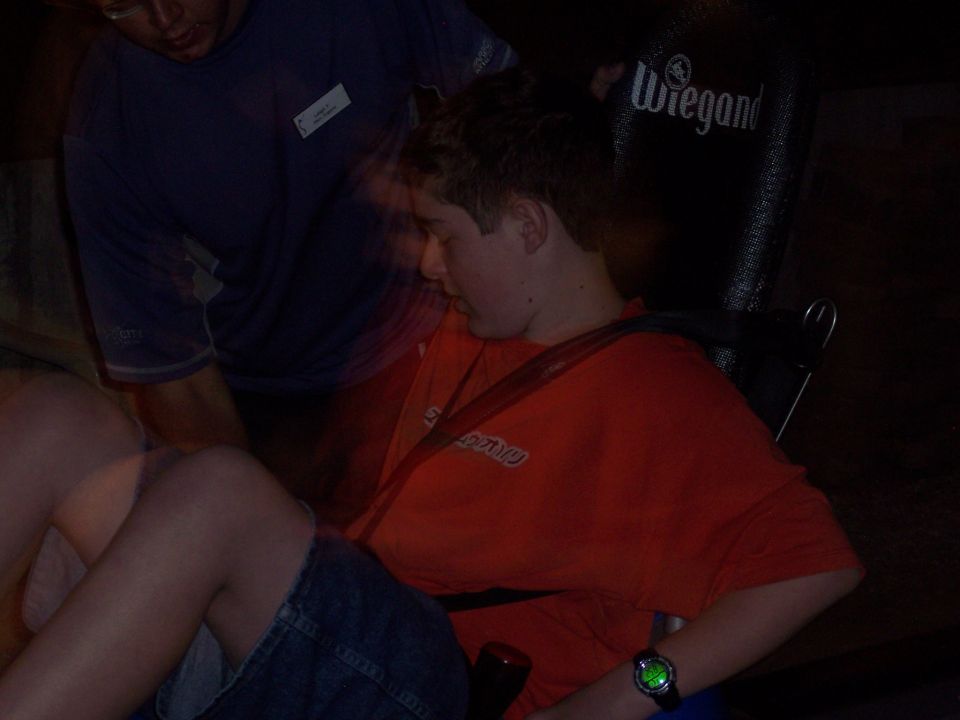 Youth in orange shirt with green-lit watch seated in a dark room, adult nearby