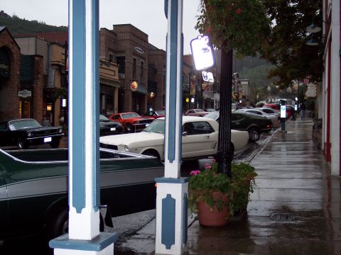 1972 Ford Ranchero - A white 1965 Ford Mustang coupe is parked on a wet street lined with shops.