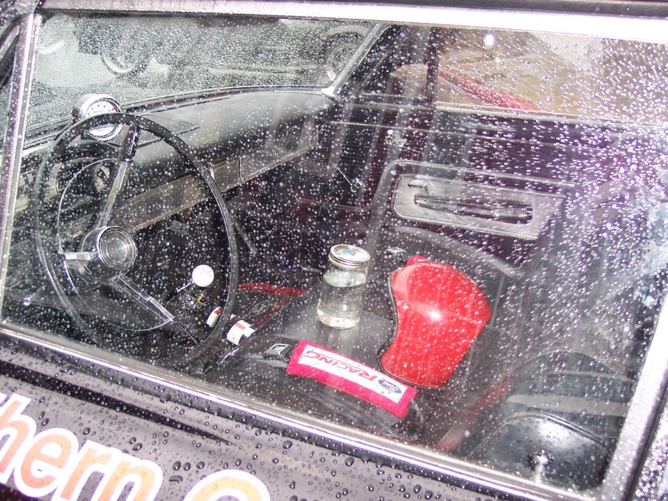 Car interior seen through wet window: black steering wheel, red shifter, glass jar, RACING pad.